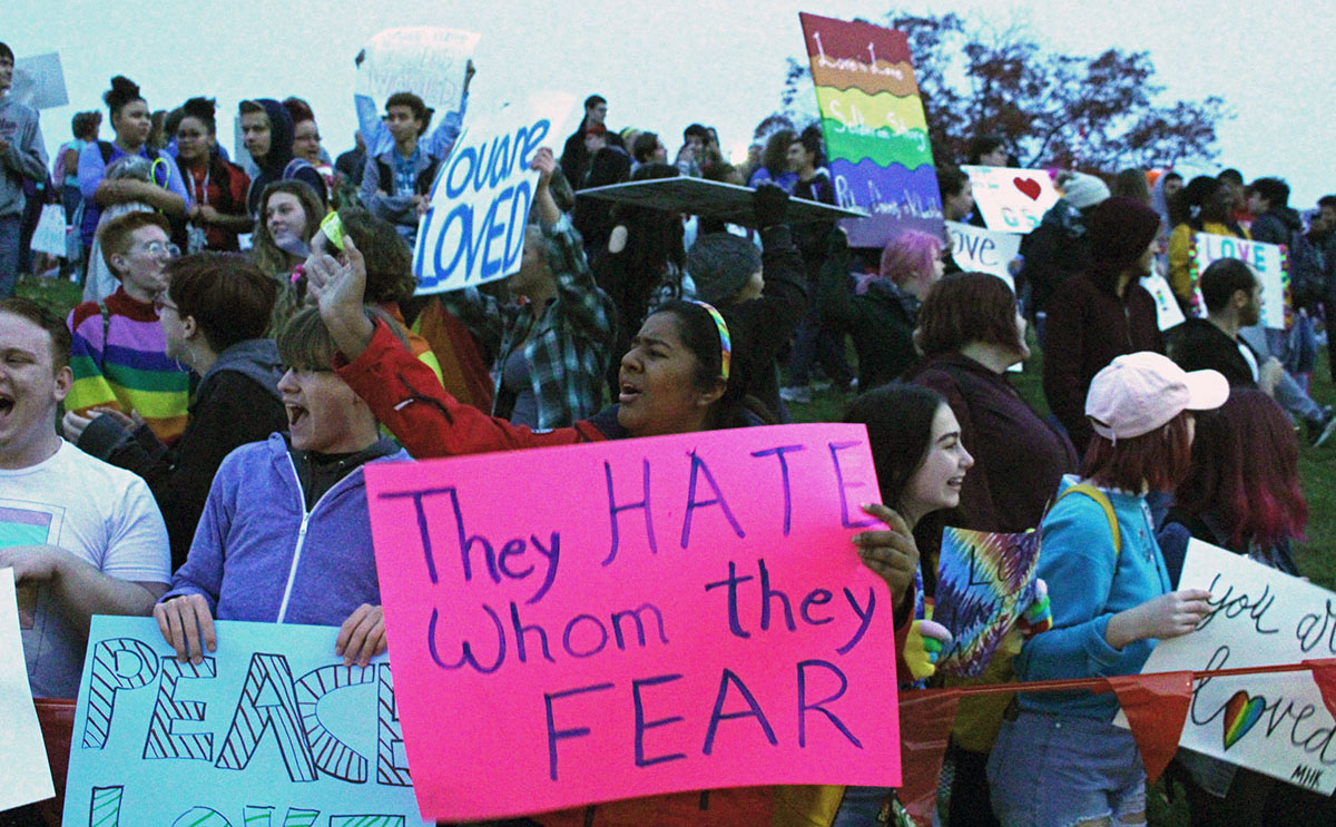 Junior Tanya Singh waves to bystanders and holds up a sign during the protest against the Westboro Baptist Church on Thursday. Students and faculty at Manhattan High School stood outside the grounds of MHS as a form of protest against the Westboro Baptist Church. Students held signs displaying their pride and support for the LGBTQ+ community like Singh's with positive messages like
