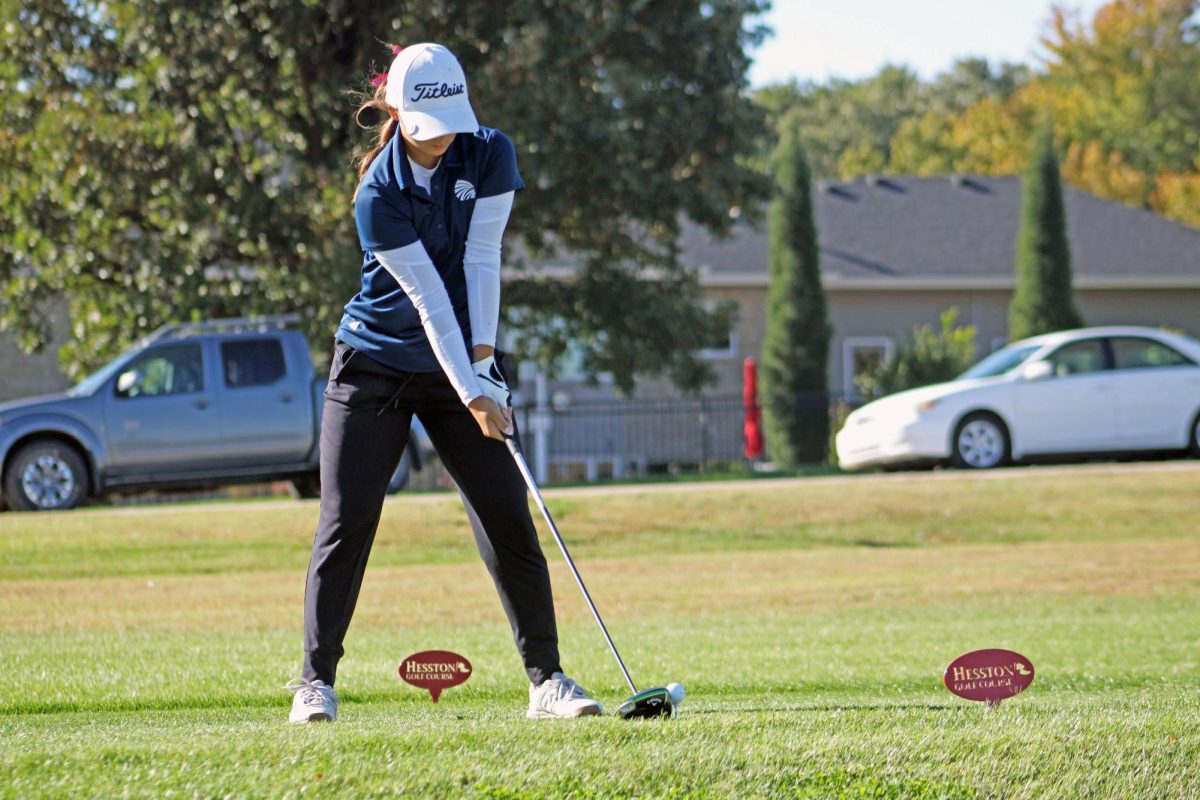 Ready to make a mark. On Oct. 20, 2025, the annual girls golf state competition kicked off in Hesston, Kansas, with Manhattan High School making the journey to tee off at 10 a.m. Senior Kat Ball positions her club against the ball, gearing up to drive it far across the golf course.