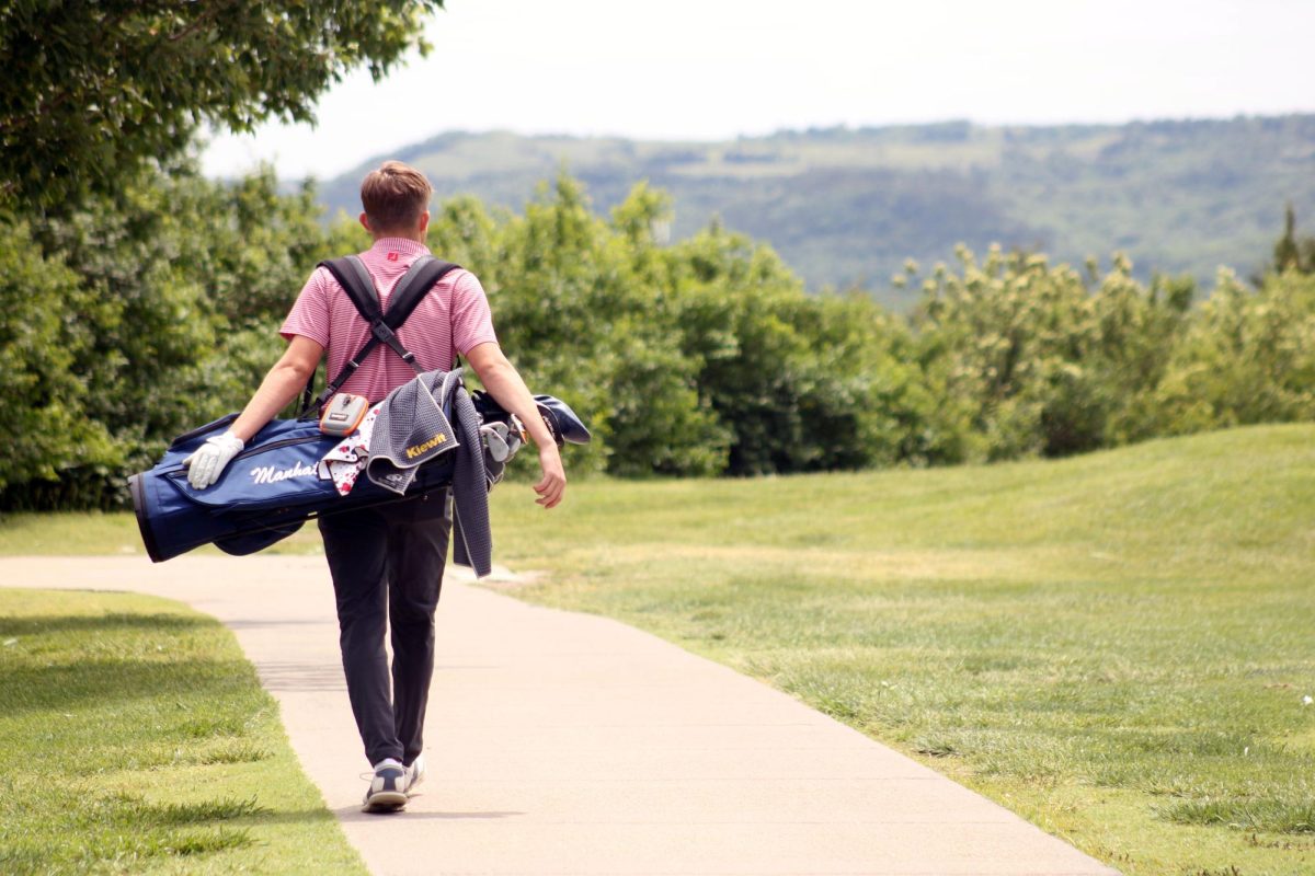 Aesthetic view. Senior Will Carpenter looks out into the green grasses of the golf course and beyond as he arrives at the club bag drop off area right next to the green with the white flag right above the hole. This was during the Substate Tournament at the Manhattan Country Club on May 20, where Carpenter played for the Manhattan High School Varsity Boys Golf Club.